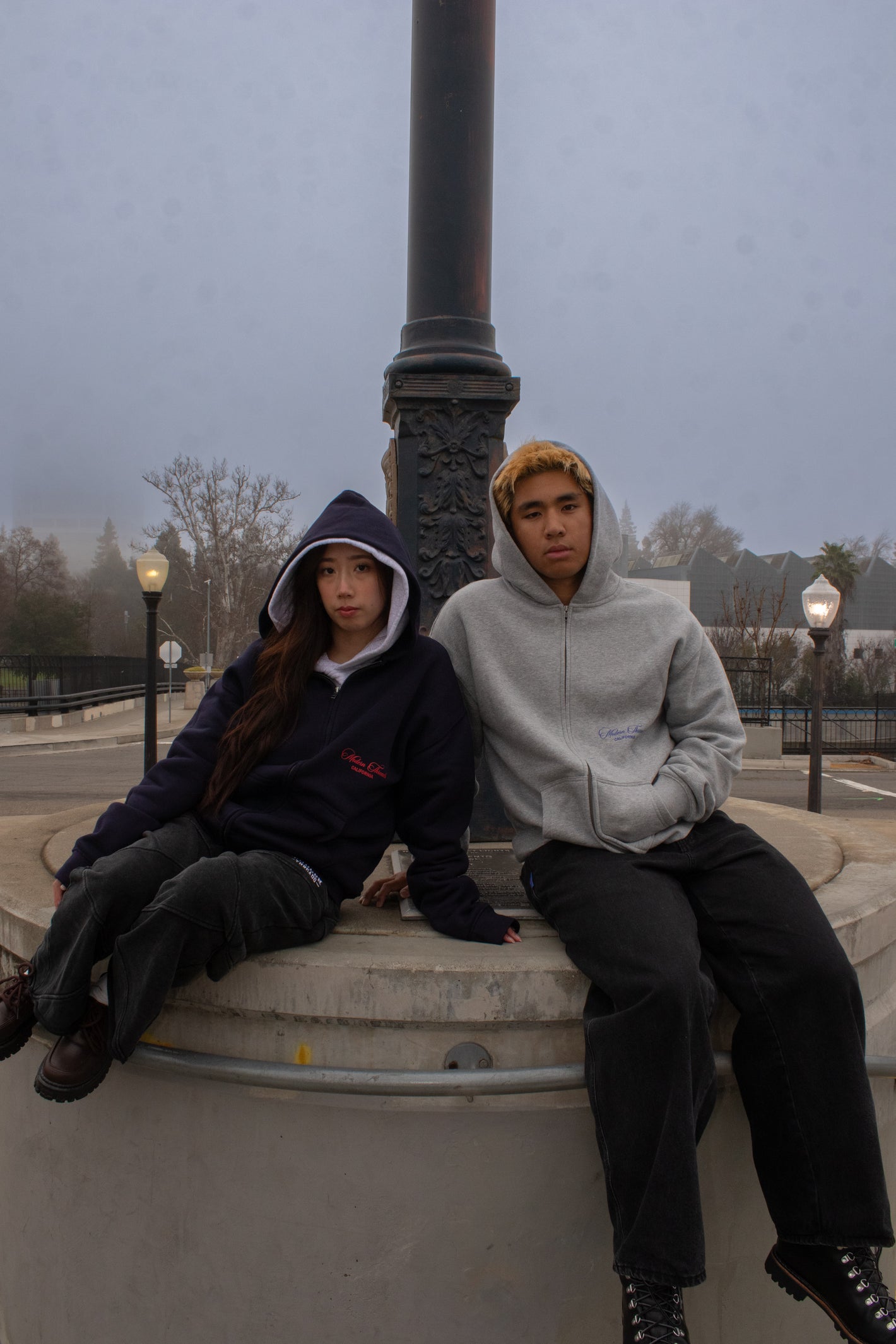Two people sitting on a concrete ledge wearing hoodies in an urban setting.